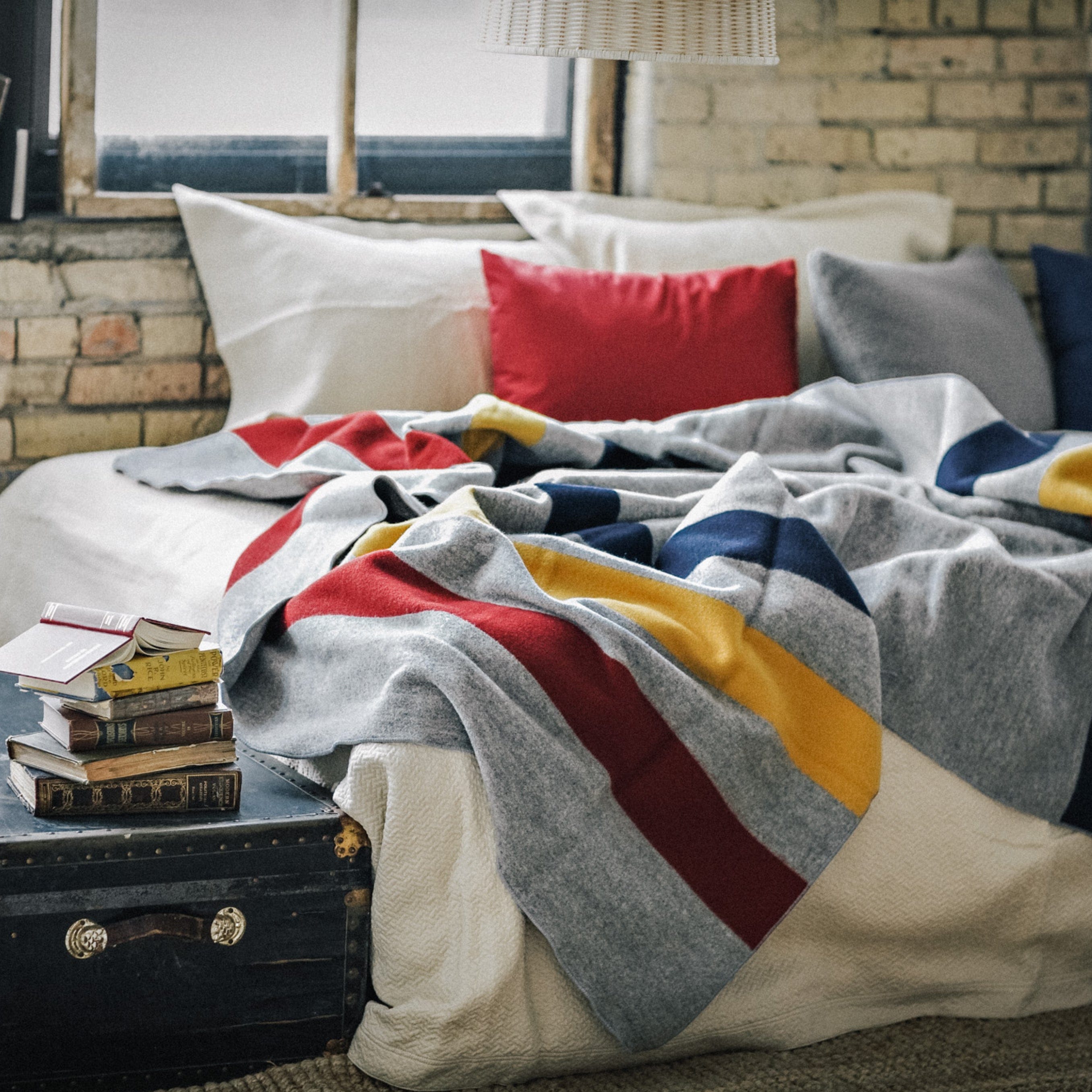 Frontier Gray heavyweight wool blanket with colorful stripes on a bed in a loft with exposed brick, a vintage trunk with books, and natural light.