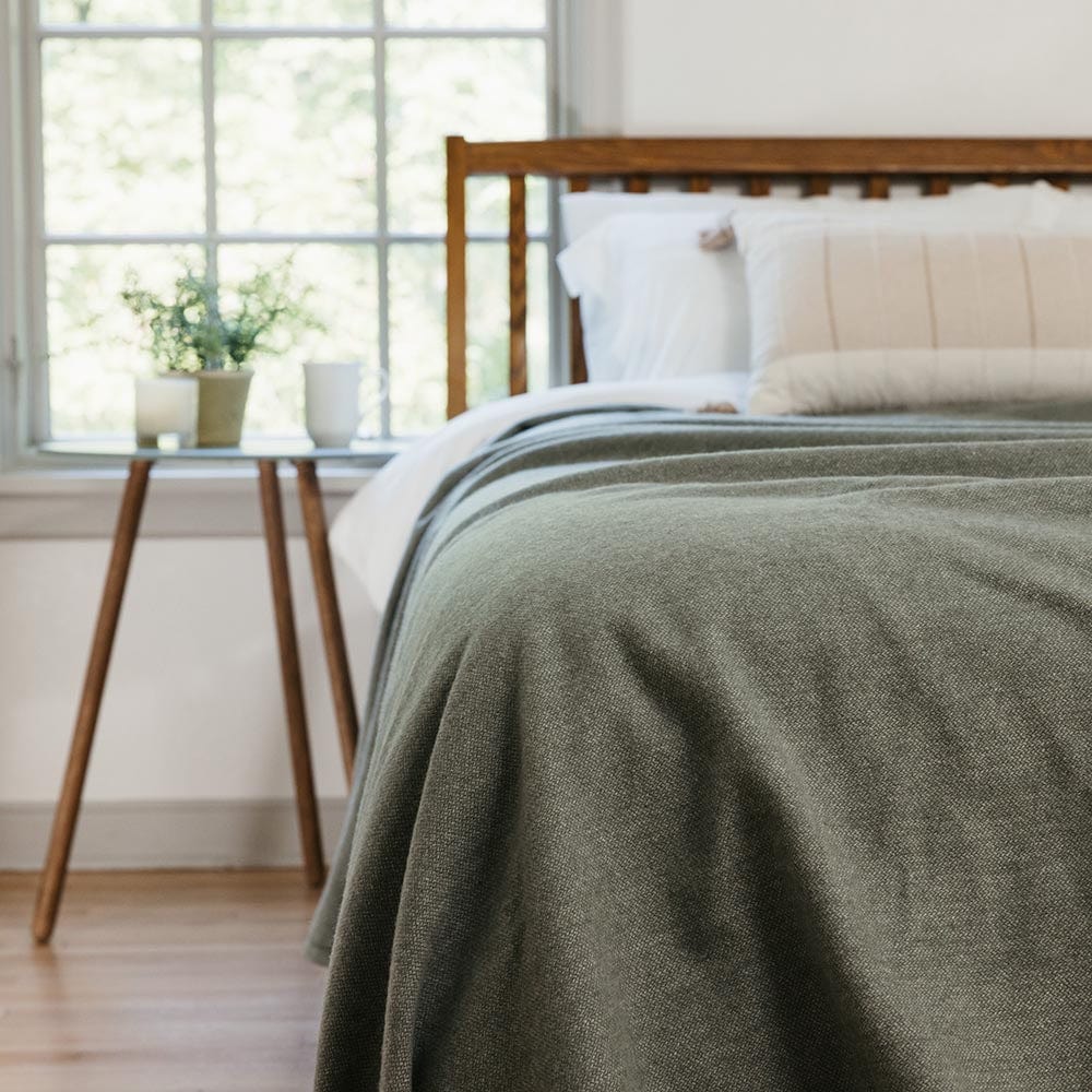 Lakefront Sage Green lightweight blanket on a bed with a wooden headboard, lots of natural light, next to a side table with a plant and window view.