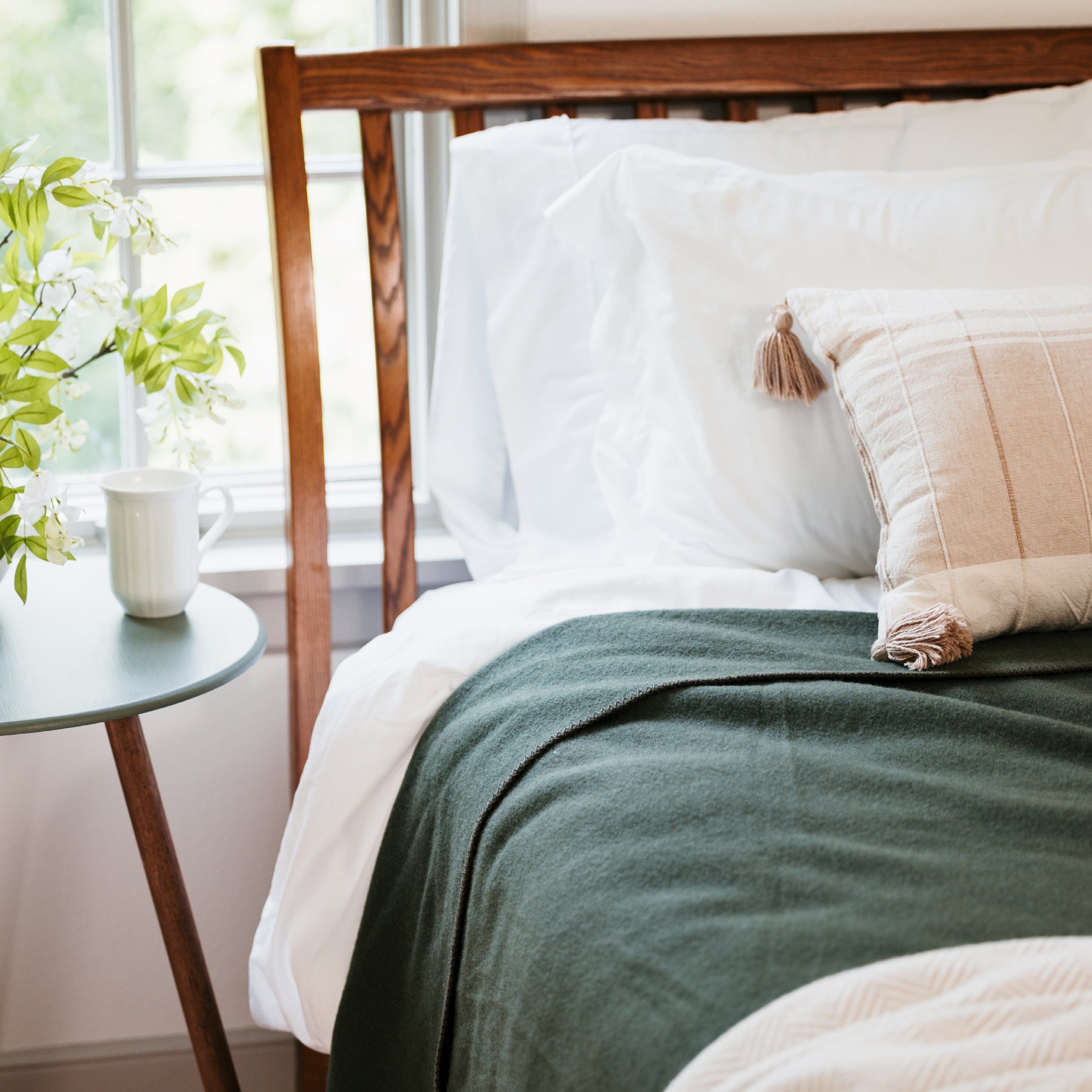 Pure and Simple Olive middleweight wool blanket on a bed with a wooden headboard, lots of natural light, next to a side table with a mug and window view.