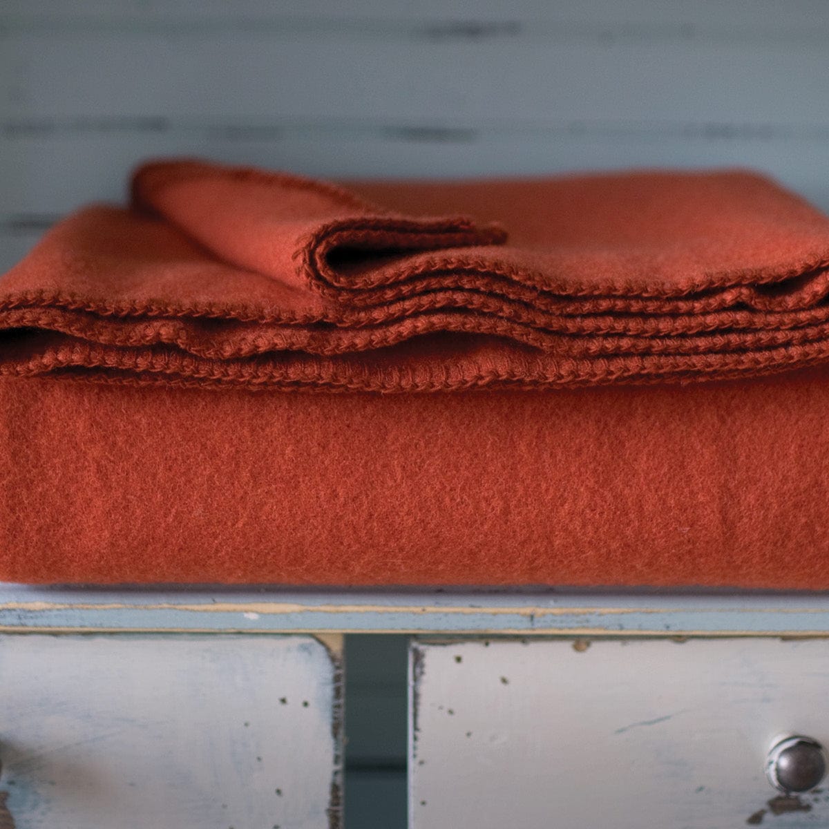 Pure and Simple Terracotta middleweight wool blanket folded on a rustic, distressed white and blue wooden cabinet in a lifestyle setting.