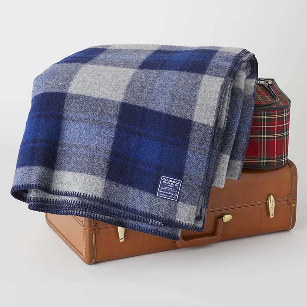 North Shore Wool Blanket in blue plaid folded over a vintage leather suitcase and a small red plaid case against a white background.