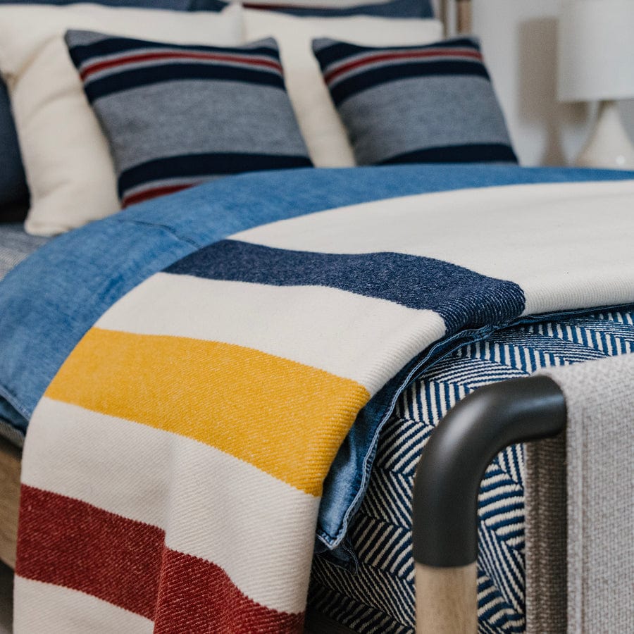 Frontier Natural heavyweight wool blanket with blue, gold, and red stripes folded on a bed, styled with coordinating blue and gray throw pillows.