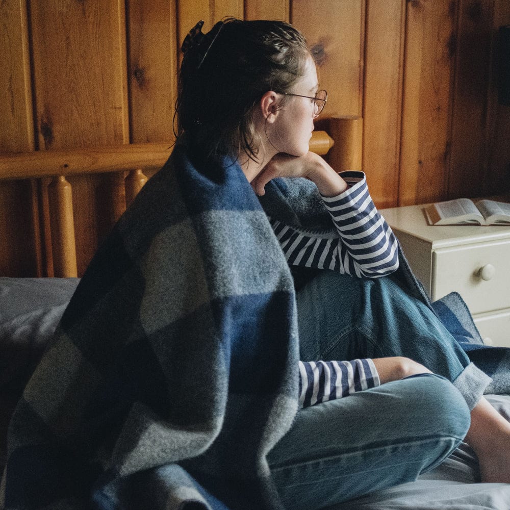 North Shore Wool Blanket in blue plaid wrapped around a woman sitting on a bed with a log headboard and wood paneled walls in a lifestyle setting.