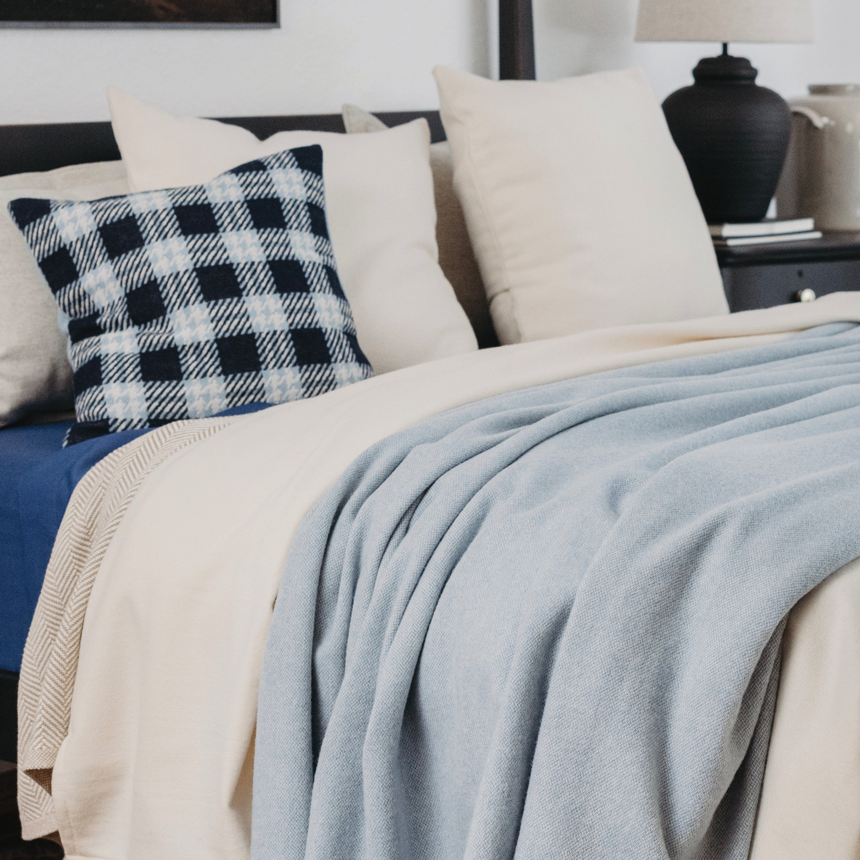 Pure & Simple Sky Blue middleweight wool blanket draped on a bed, styled with white pillows and a dark blue and white gingham check throw pillow.