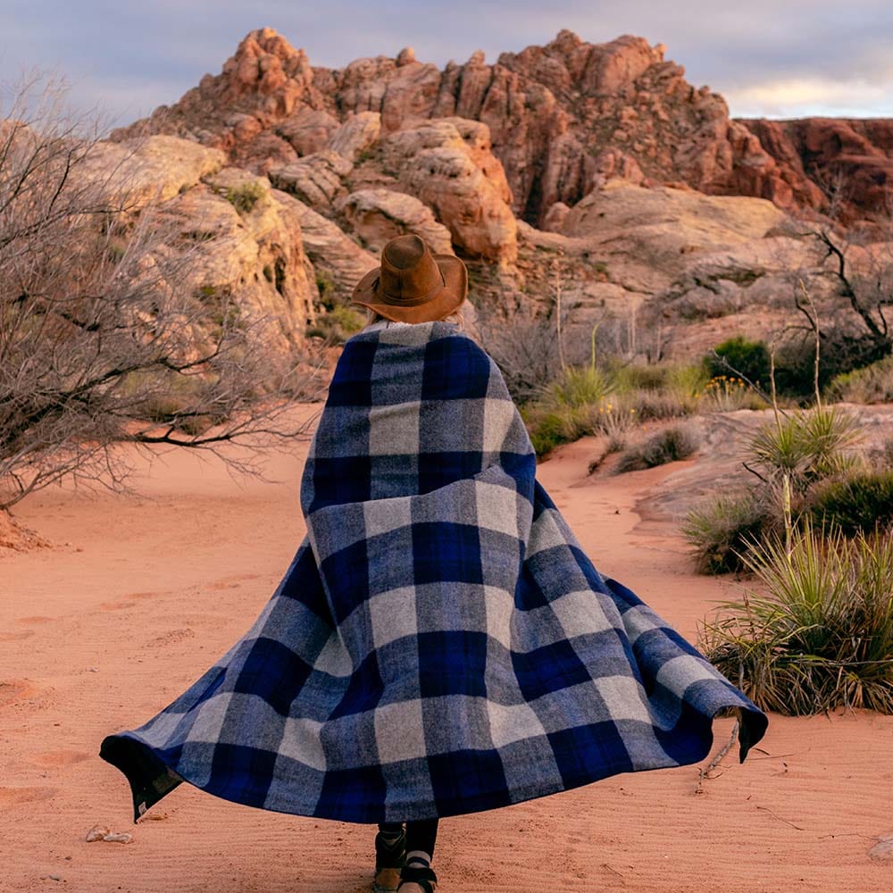 North Shore Wool Blanket in blue plaid wrapped like a cape around a person walking through a desert landscape with rocky formations and sand.
