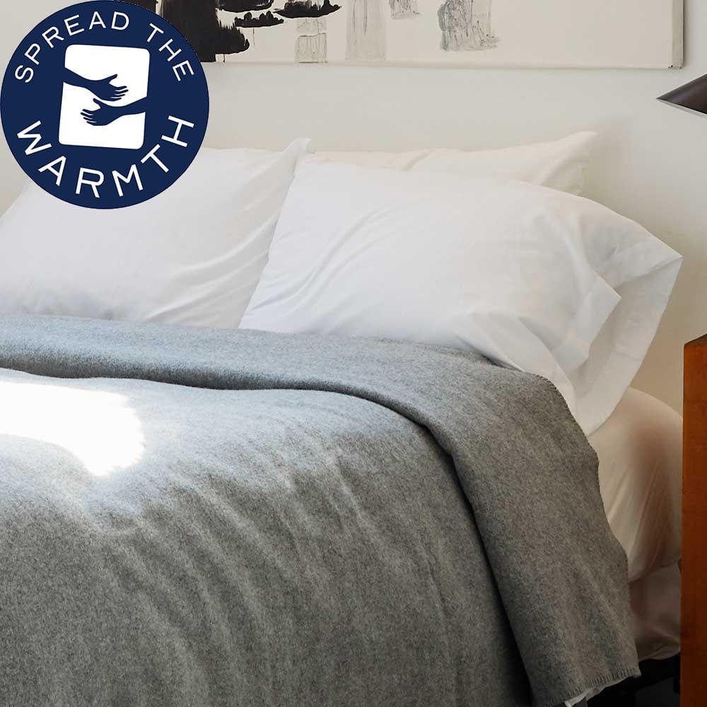 Pure and Simple Heather Gray middleweight wool blanket on a bed with white pillows, styled next to a wooden nightstand in a bright room.